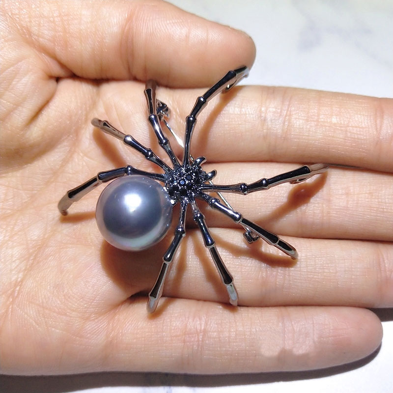 Broche araignée noire avec perle grise ronde tenue dans une main, détail des pattes fines et finition soignée