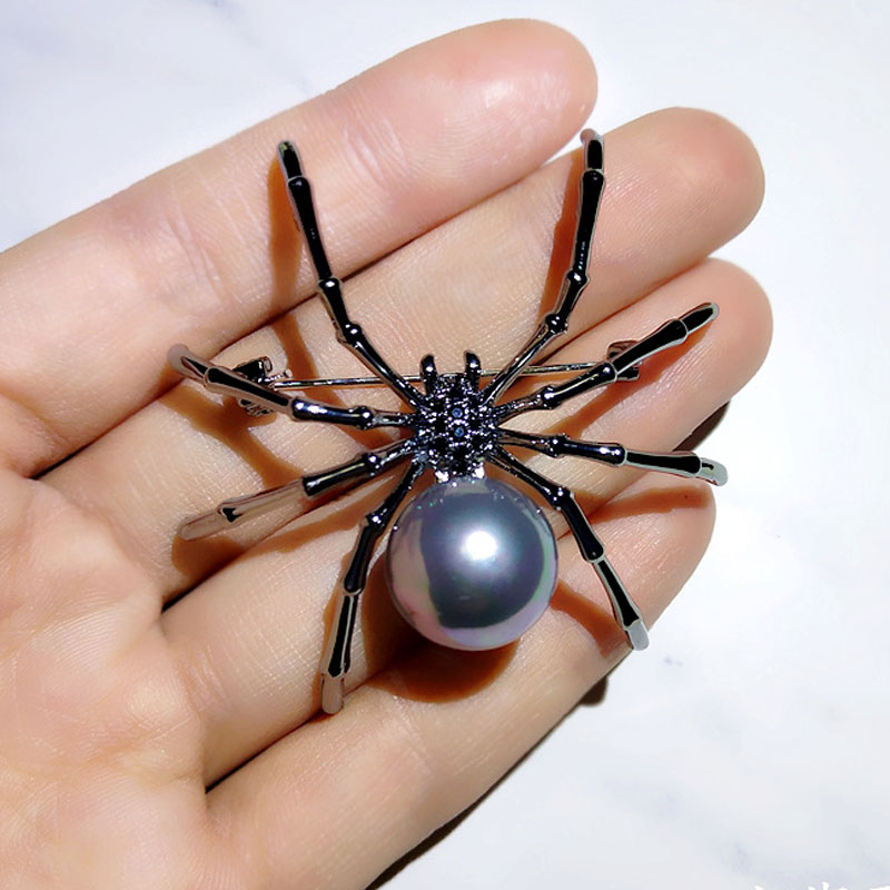 Broche araignée noire avec perle grise tenue dans une main, détail des pattes fines et finition soignée