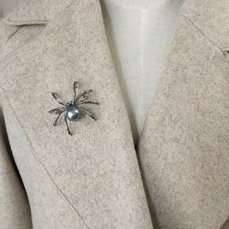 Broche araignée en métal argenté avec perle grise centrale portée sur un manteau beige en laine
