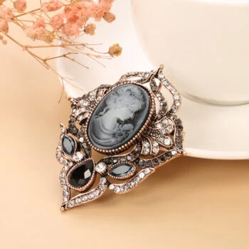 Broche camée femme en métal doré avec cabochon ovale noir et blanc représentant un profil féminin, ornée de strass et de pierres noires, détail raffiné et design élégant