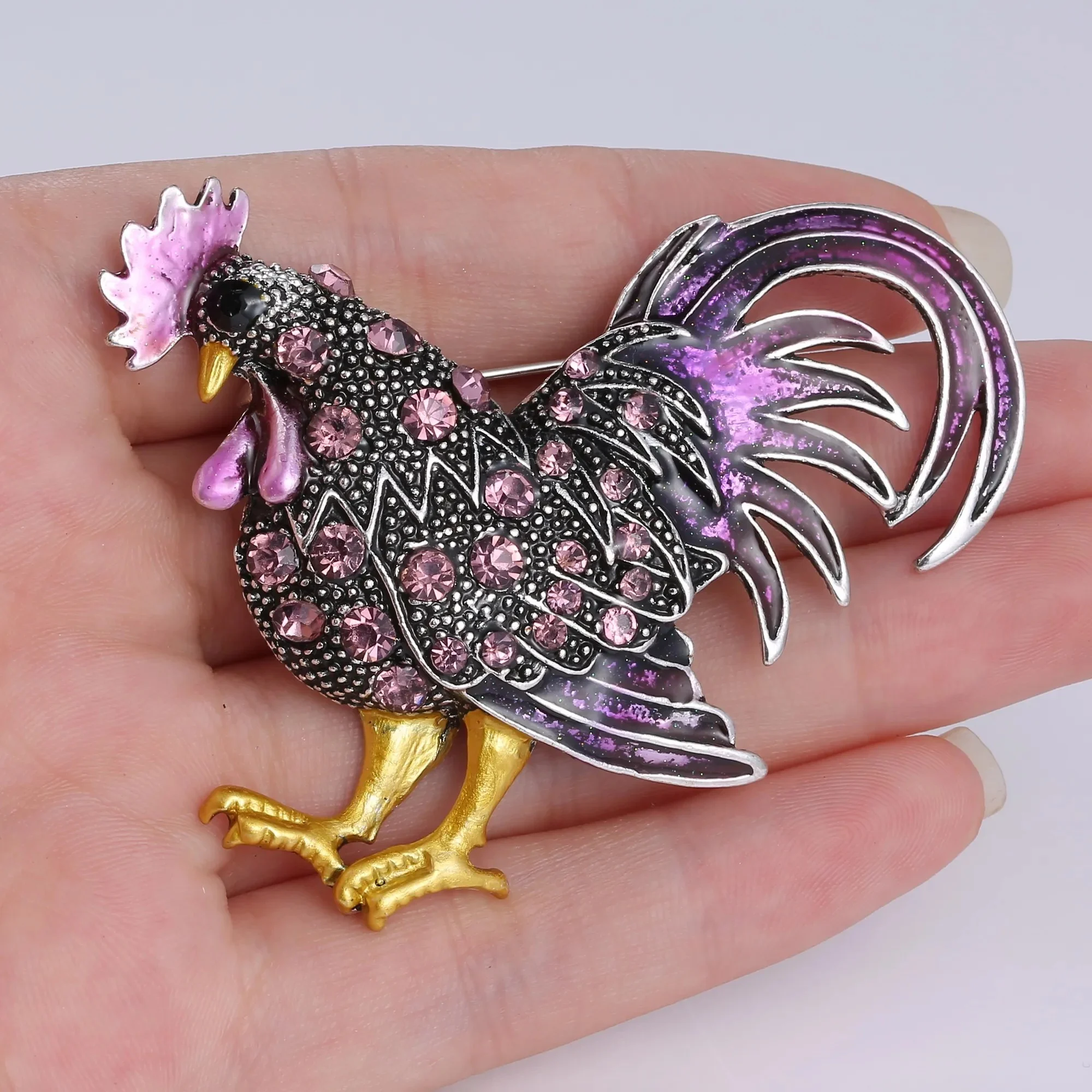 Broche Coq Fantaisie en métal avec motifs floraux roses et détails violets, pattes dorées, tenue dans une main