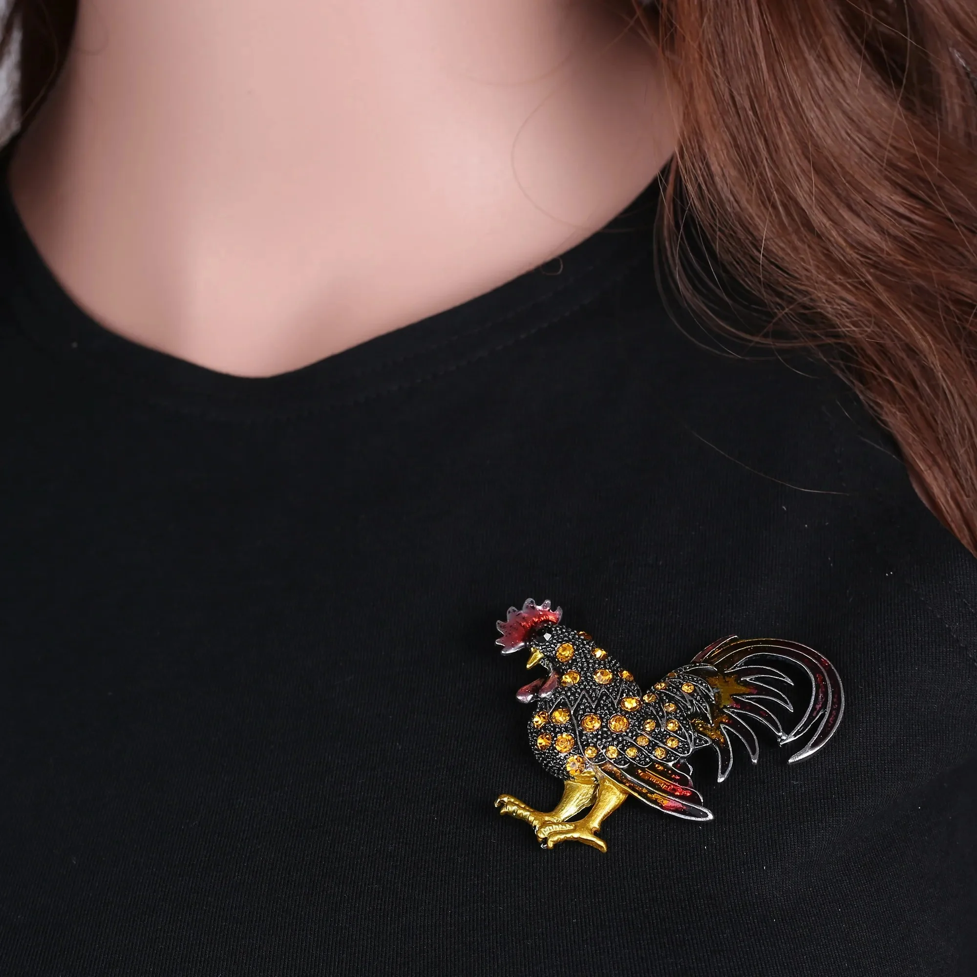Broche coq fantaisie en métal doré avec strass jaunes et rouges portée sur un vêtement noir, vue rapprochée angle légèrement de côté