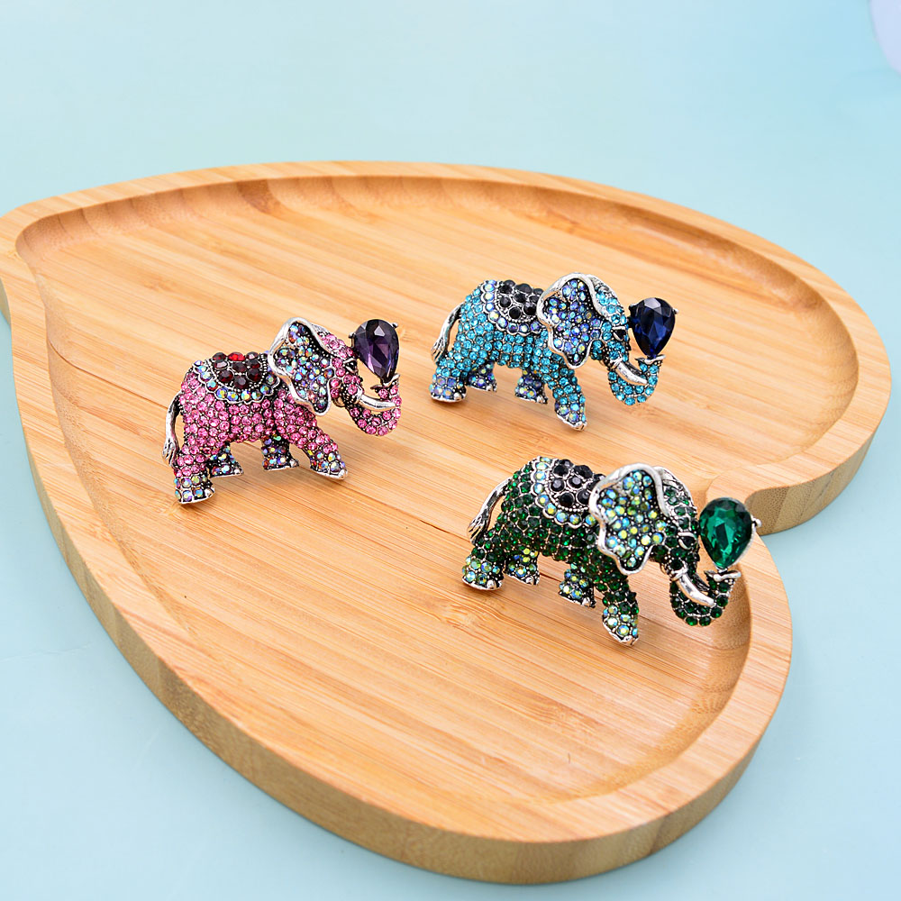 Broche éléphant fantaisie en métal avec strass multicolores rose, bleu et vert posée sur plateau en bois en forme de cœur, détail soigné des motifs et finition élégante