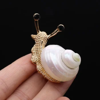 Broche escargot en métal doré avec coquille nacrée blanche tenue entre les doigts sur fond noir