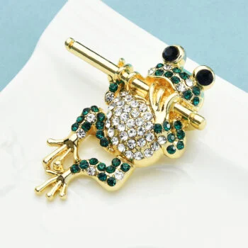 Broche grenouille dorée à la flûte ornée de strass verts et blancs, détail soigné sur fond blanc