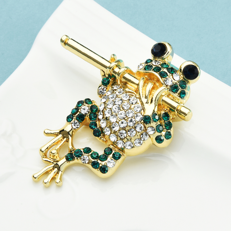 Broche grenouille dorée à la flûte ornée de strass verts et blancs, détail soigné sur fond blanc