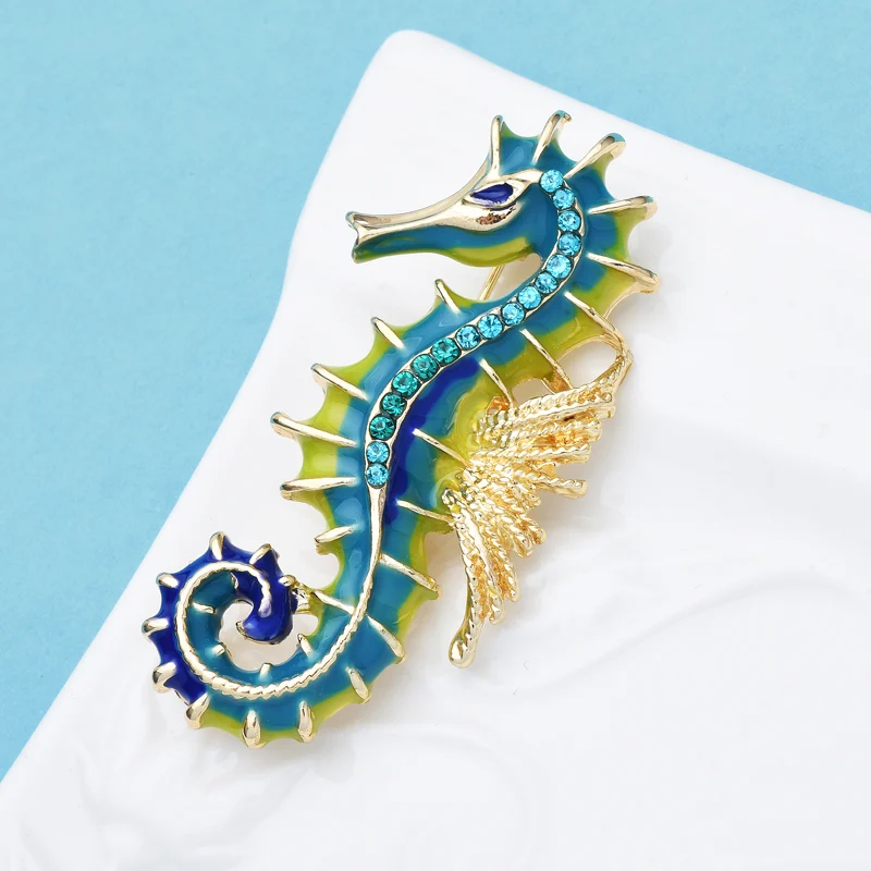Broche hippocampe en métal doré avec émail bleu et vert, ornée de petits strass bleus sur le corps