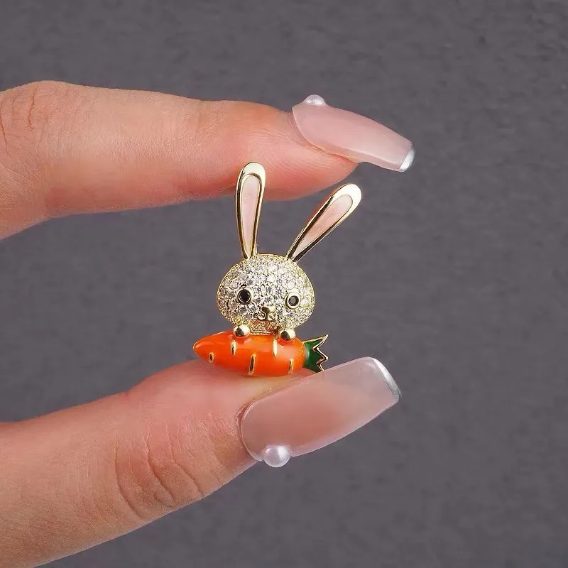 Broche lapin et carotte strass tenue entre deux doigts, détail de la tête en strass et de la carotte orange avec feuilles vertes