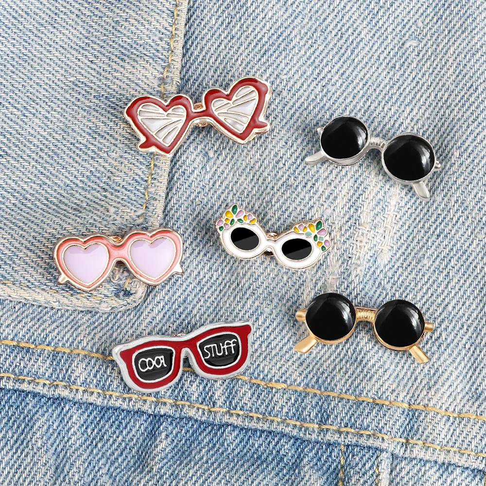 Broche lunettes en émail avec plusieurs modèles colorés sur tissu jean, incluant lunettes cœur rouges, lunettes cœur roses, lunettes rondes noires, lunettes blanches à motifs colorés et lunettes rectangulaires noires avec texte