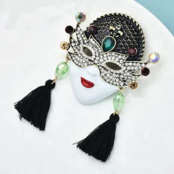 Broche Masque Vénitien en métal noir et argenté avec strass blancs, pierres vertes et bordeaux, pompons noirs et visage blanc au maquillage rouge