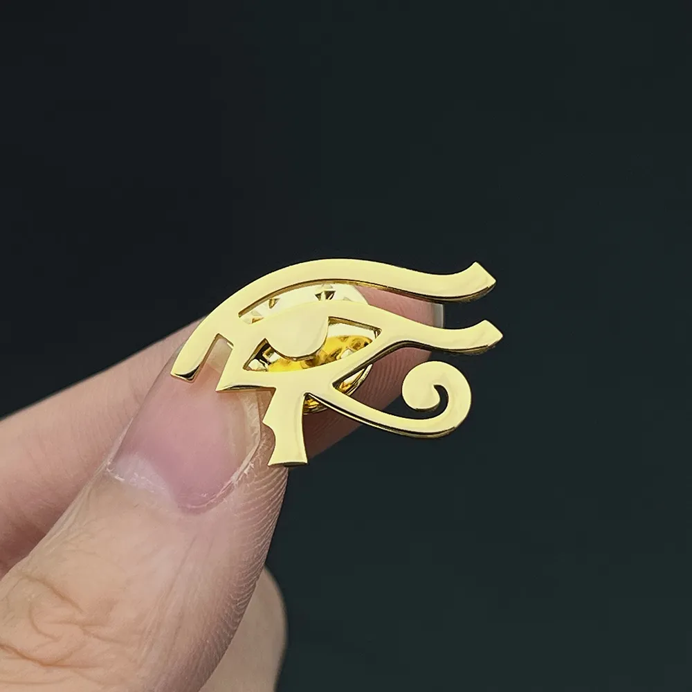 Broche Œil d'Horus en métal doré tenue entre deux doigts, détail du motif égyptien stylisé