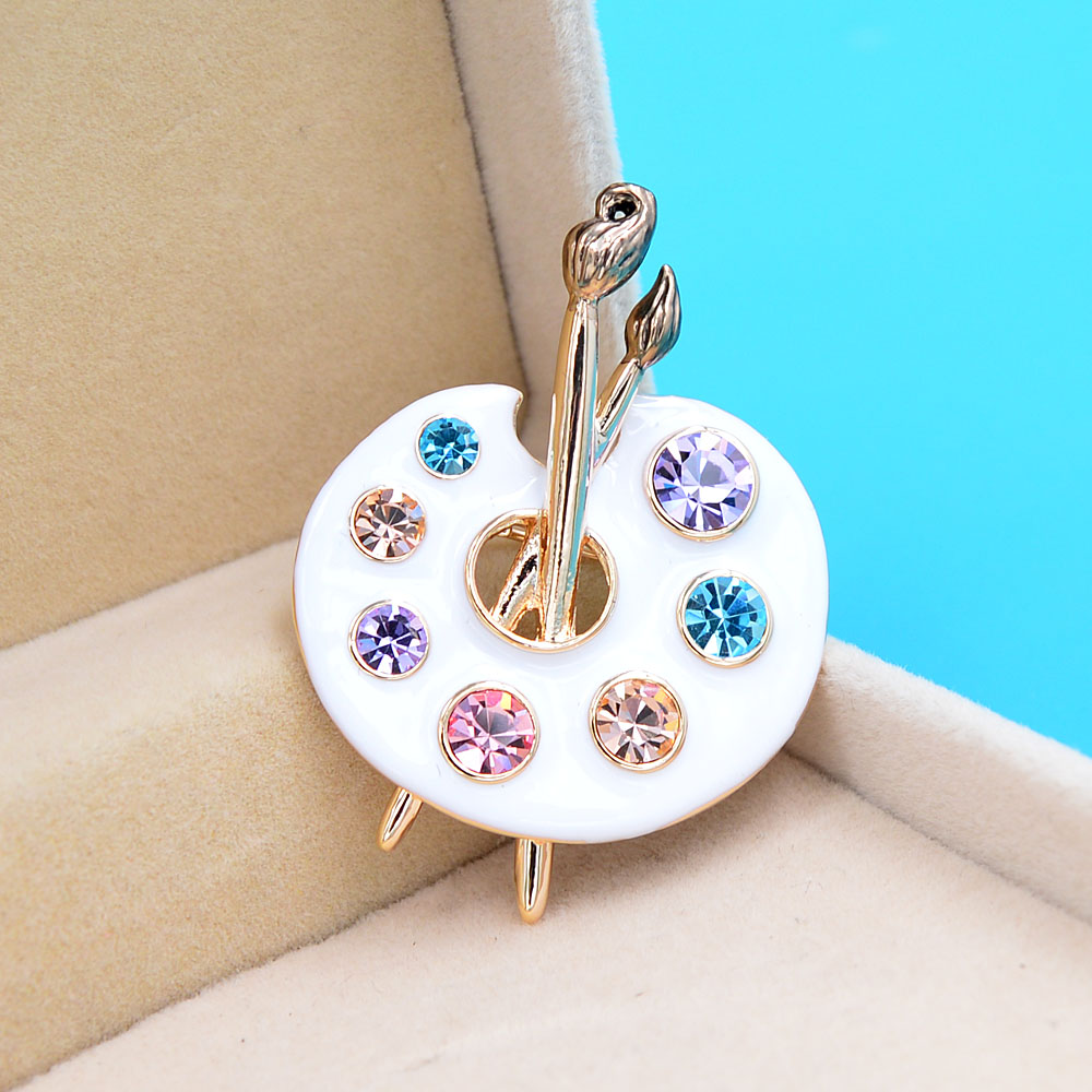 Broche palette de peintre blanche en métal doré avec strass colorés bleu, violet et rose, détail du pinceau en position verticale