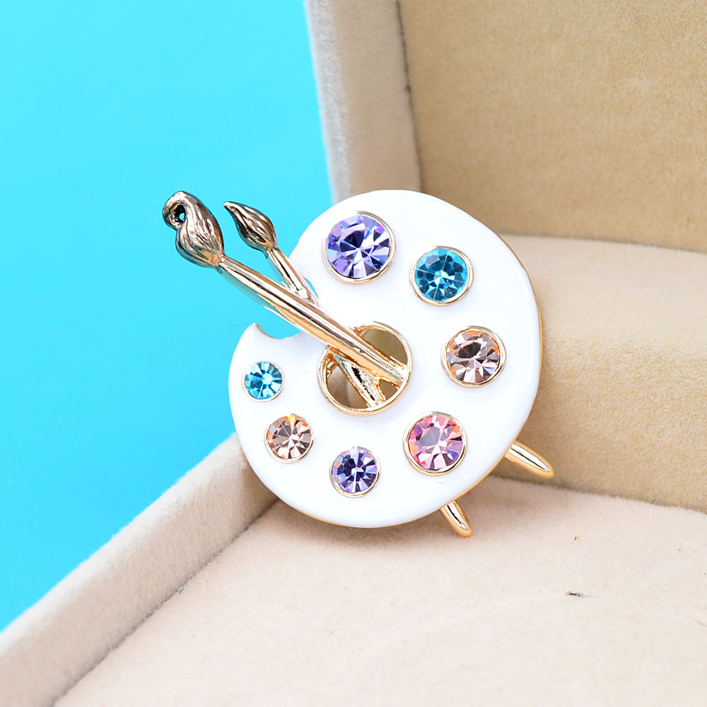 Broche palette de peintre blanche en métal doré avec strass colorés et pinceaux détaillés