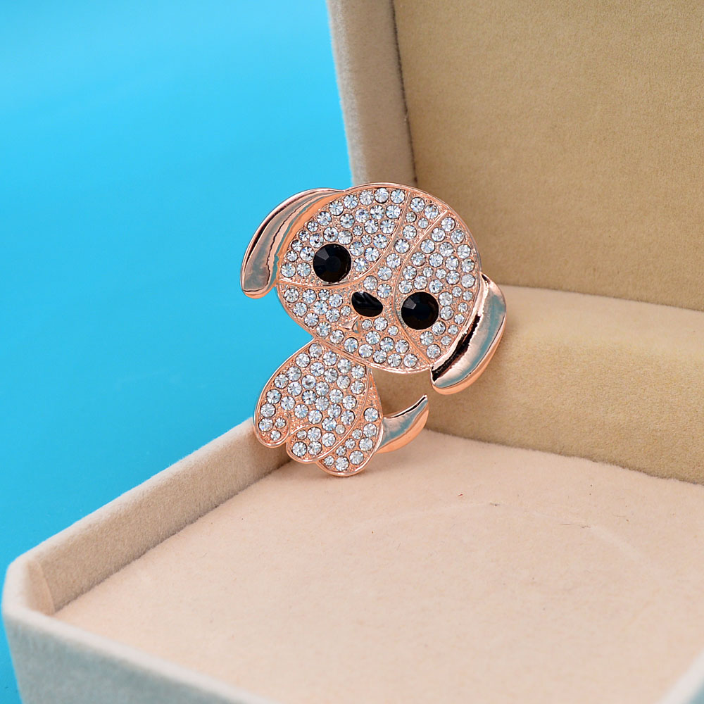 Broche petit chien en métal doré rose avec strass blancs et yeux noirs, vue en léger angle dans un écrin beige