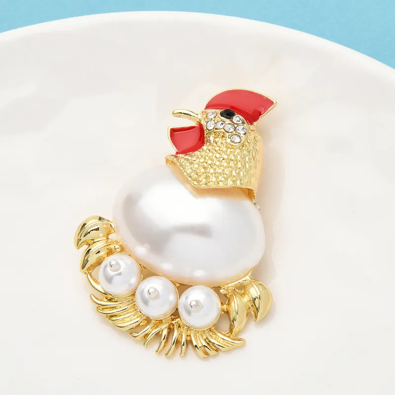 Broche poule dorée avec tête rouge ornée de strass, corps en perle blanche et base décorée de perles nacrées sur fond bleu clair