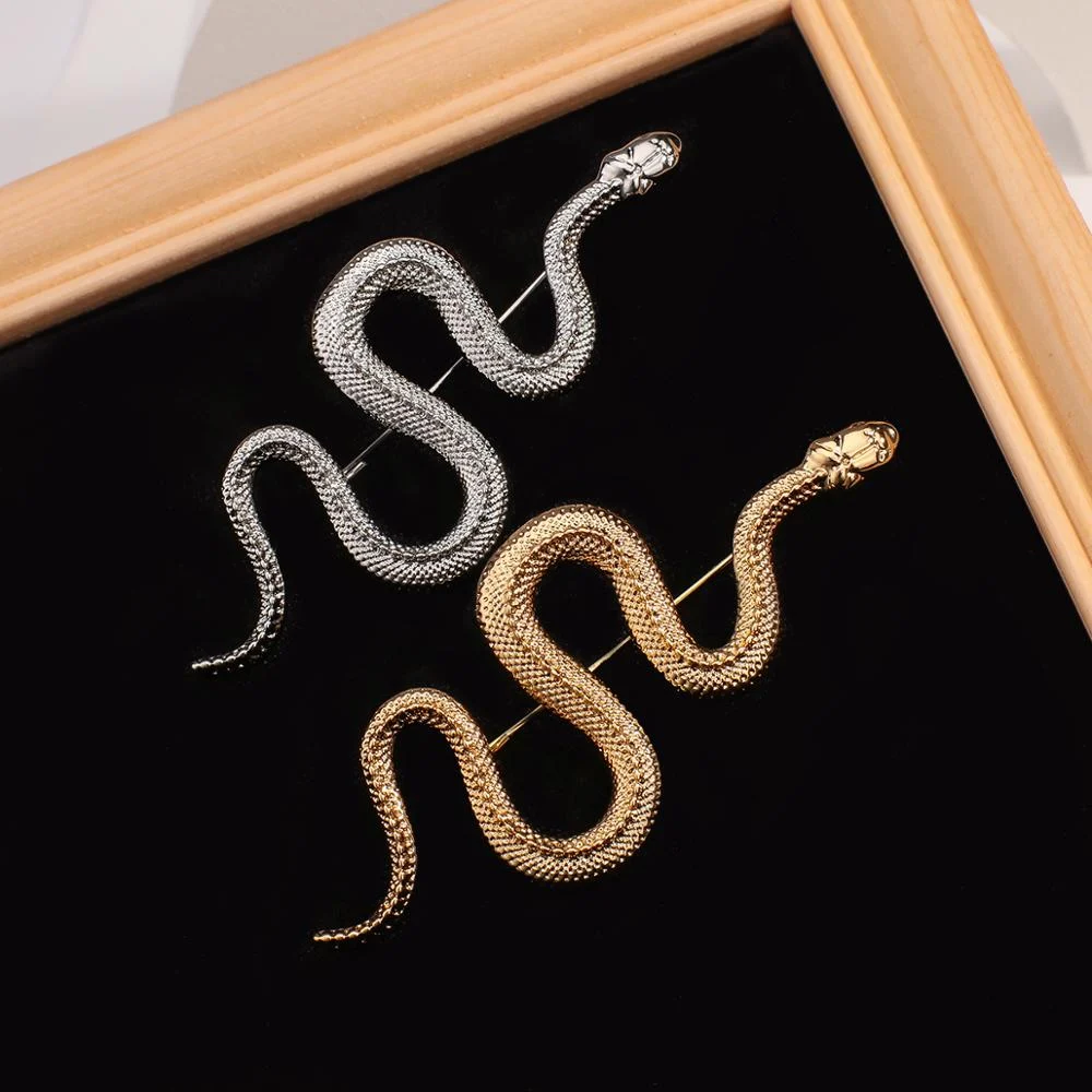 Broche serpent en métal doré et argenté avec finition texturée posée sur fond noir dans un cadre en bois clair