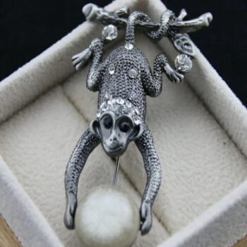 Broche singe en métal argenté avec finition texturée et yeux noirs, tenant une boule blanche duveteuse, présentée dans un écrin beige