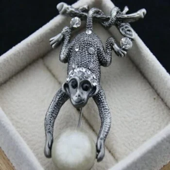 Broche singe en métal argenté avec finition texturée et yeux noirs, tenant une boule blanche duveteuse, présentée dans un écrin beige