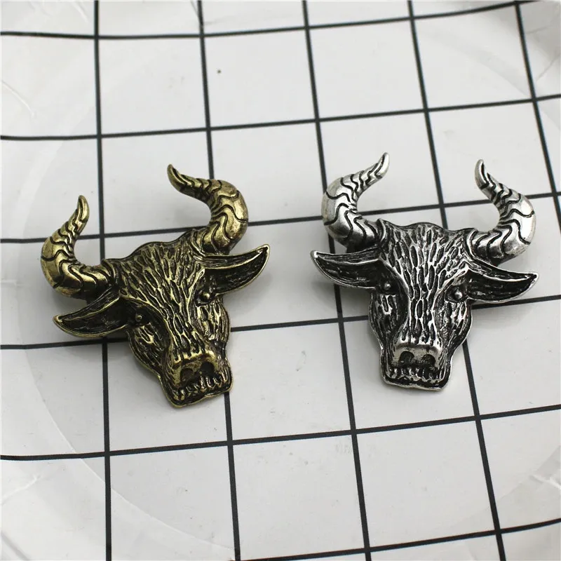 Broche tête de taureau en métal, une en finition dorée vieillie et l'autre en finition argentée, posées sur un fond quadrillé blanc et noir