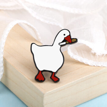 Petit pin's oie blanche en métal émaillé avec bec et pattes rouges posé sur un coin de bois clair, fond tissu blanc transparent