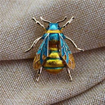 Petite broche abeille en métal doré avec émail bleu et jaune, détail soigné des ailes et du corps, posée sur tissu beige