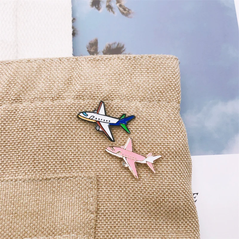 Pin's avion en métal émaillé, modèle double avec un avion blanc et bleu et un avion rose, posé sur tissu beige