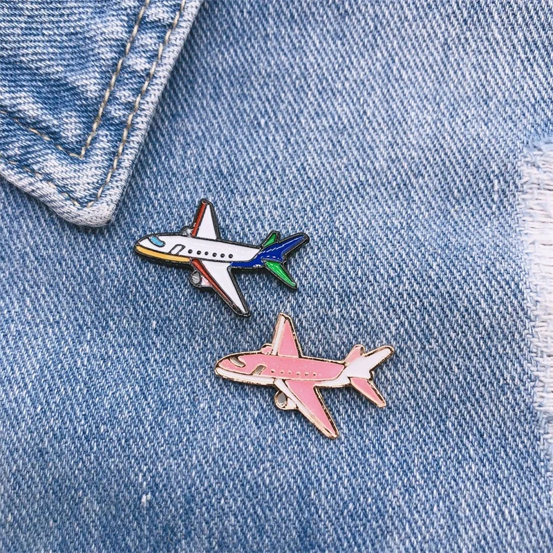 Pin's avion en métal émaillé, un modèle blanc et bleu avec détails verts et un modèle rose, posé sur tissu jean bleu