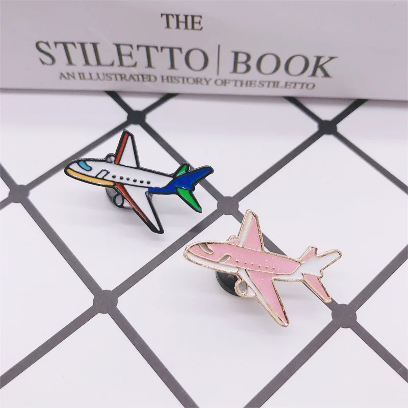 Pin's avion rose en métal avec détails en émail, posé sur surface à motif quadrillé avec un pin's avion blanc, bleu et vert en arrière-plan