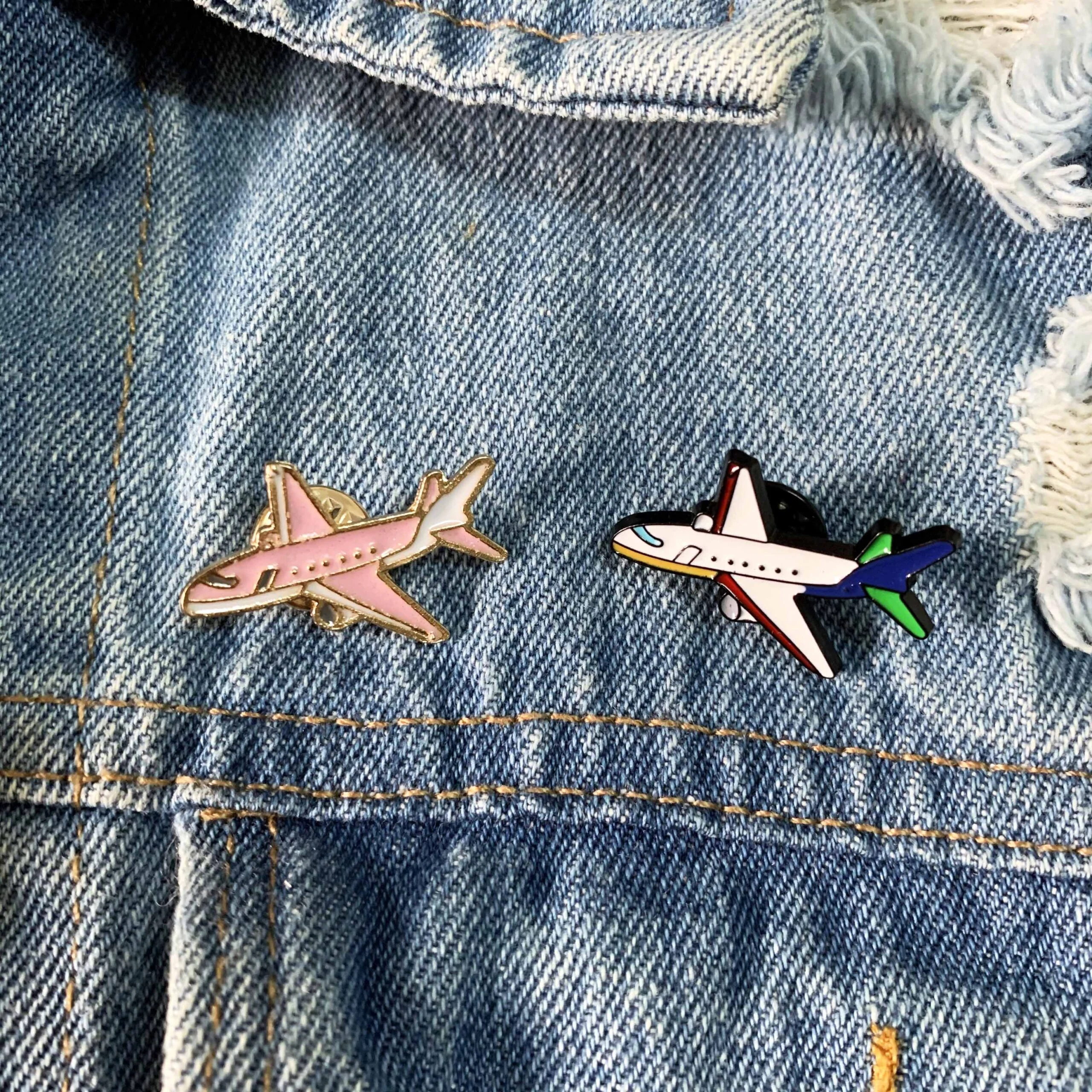 Pin's avion en métal émaillé rose avec détails dorés, posé sur un tissu en jean bleu délavé