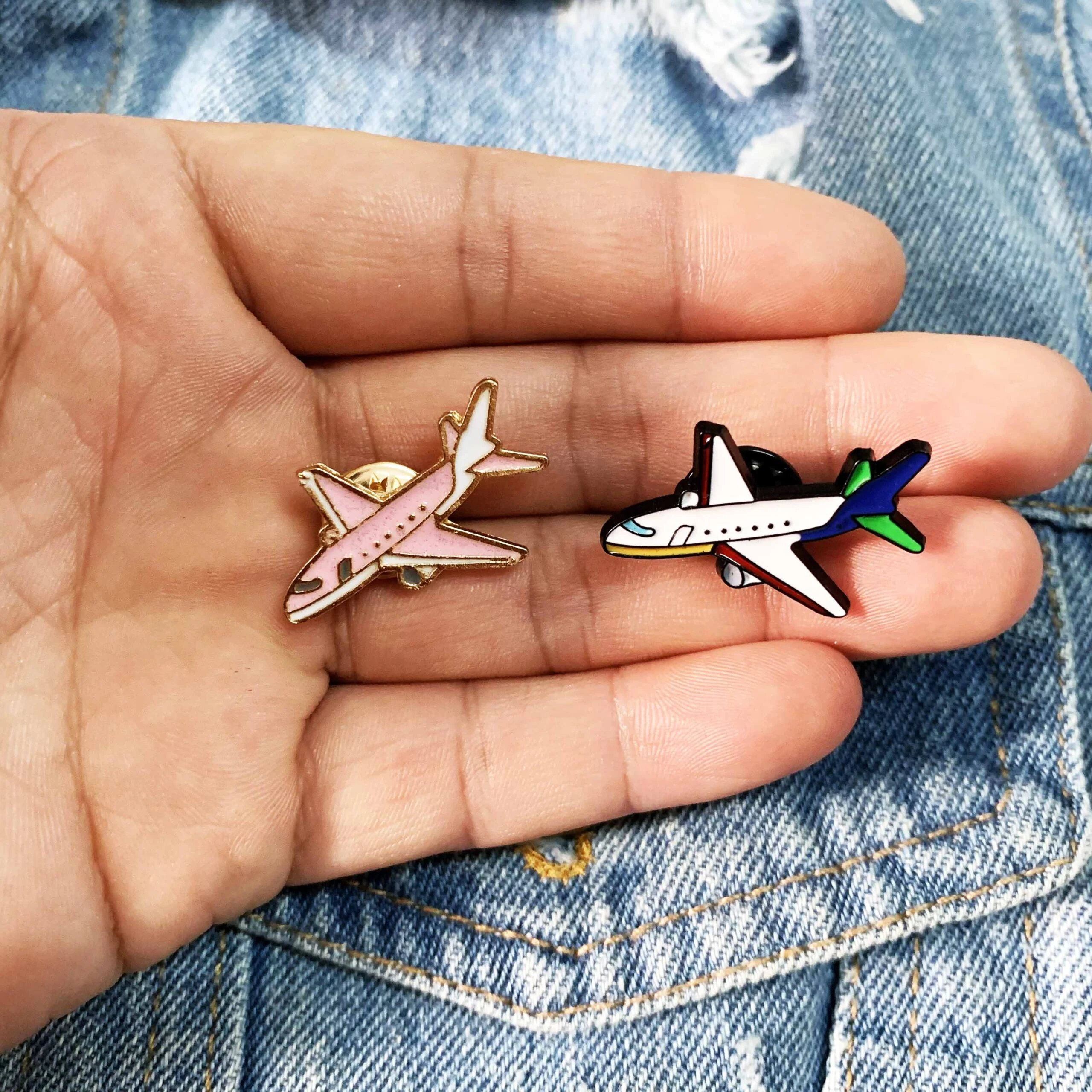 Pin's avion en métal émaillé, modèle rose pâle avec détails dorés, tenu dans une main sur un fond de veste en jean