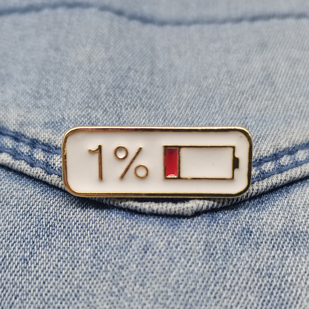 Pin's barre d'énergie en métal doré avec fond blanc et indicateur de batterie à 1% rouge, posé sur tissu jean bleu
