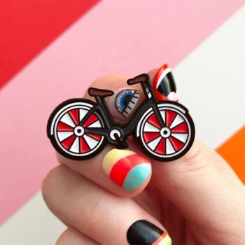Pin's bicyclette en métal avec roues rouges et blanches, cadre marron, détail bleu sur le cadre, tenue par une main avec ongles colorés
