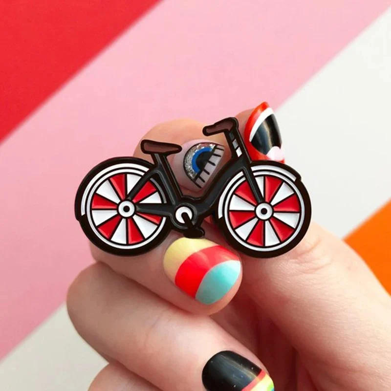 Pin's bicyclette en métal avec roues rouges et blanches, cadre marron, détail bleu sur le cadre, tenue par une main avec ongles colorés