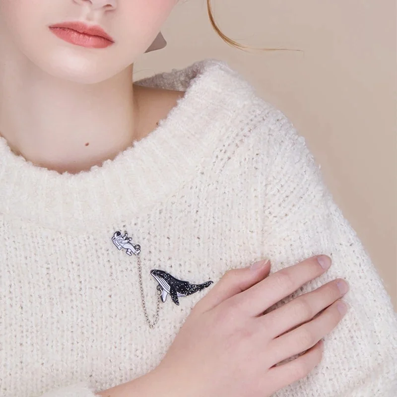 Pin's à chaîne argentée avec motif astronaute et baleine en métal émaillé noir et blanc porté sur pull blanc en maille
