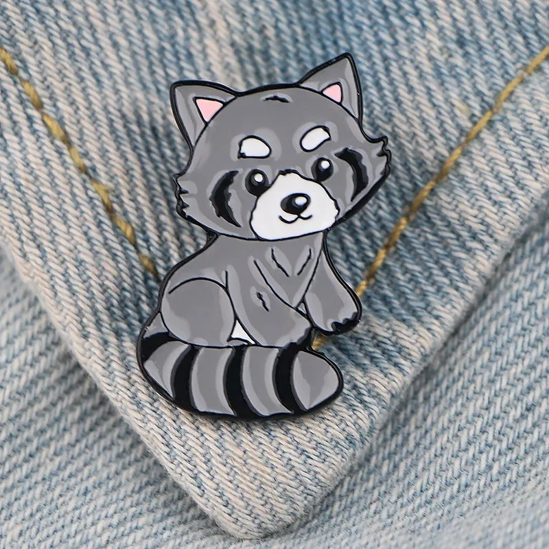 Pin's raton laveur gris en émail posé sur tissu jean clair, motif animal stylisé avec détails soignés