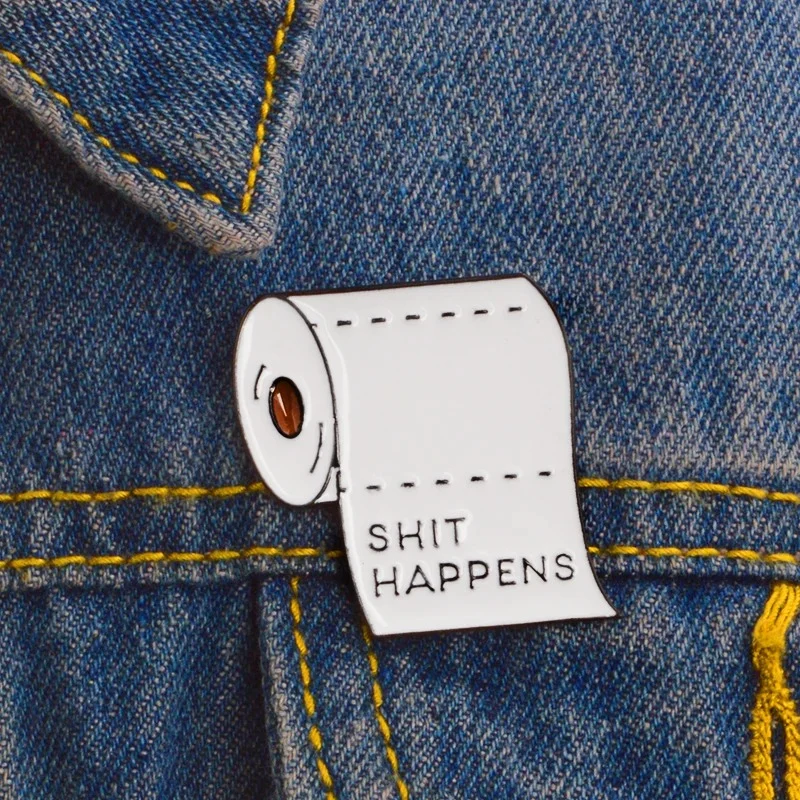 Pin's rouleau de papier toilette blanc avec inscription noire "SHIT HAPPENS" épinglé sur tissu jean bleu avec couture jaune