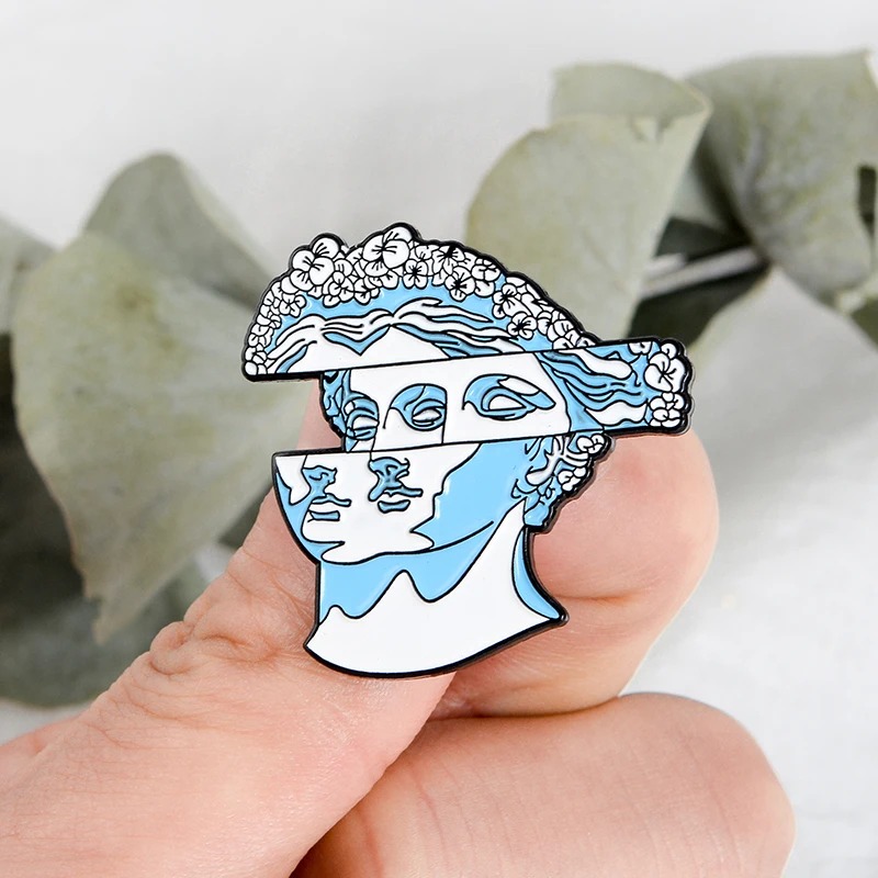Pin's visage statue grecque artistique en métal émaillé bleu et blanc avec détails floraux sur la tête, vue de face tenue entre les doigts