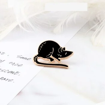 Pin's souris noire en métal avec détails dorés posée sur une surface claire à côté d'une plume blanche